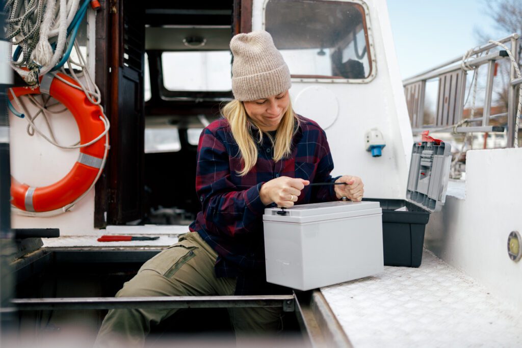 A focused fisherwoman in a beanie and plaid shirt is engaged in changing the battery on her boat, ensuring her equipment is powered and ready for a day at sea, with safety gear like a lifebuoy in the background.