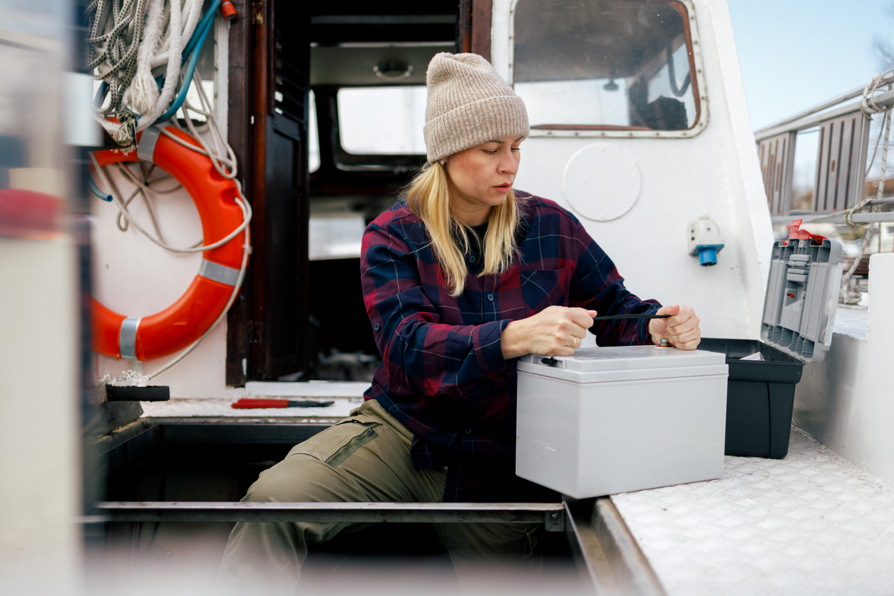 A focused fisherwoman in a beanie and plaid shirt is engaged in changing the battery on her boat, ensuring her equipment is powered and ready for a day at sea, with safety gear like a lifebuoy in the background.